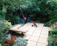 Childrens garden with swings with bark beneath, blue trellis screens, paving, wooden seat, Phormium and Ficus - fig tree