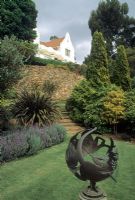 Metal ornament on plinth as focal point in lawn and steps up through borders of evergreen conifers and Phormium 