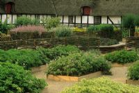 Rasied Herb borders edged with wooden planks in front of thatched house. Woven willow fencing used as boundary and divider. 