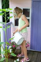 Young girl watering containers of Impatiens - Busy Lizzies with watering can outside of painted wendy house