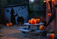 Halloween display in gravel courtyard with blue table, pumpkins, besom broom, blue shed with witch and couldron silhouette cut from pvc pondliner