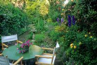 Small town cottage style garden with table and directors chairs on brick patio. Delphinium, Campanula, Alchemilla mollis and Roses in border. Impatiens in pot on table - London