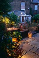 Small town back garden at night with paved patio area with table and chairs.  
Railway sleeper raised water feature and container of Oleander - London 