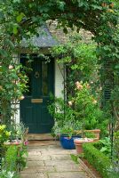 View through an arch to a front door of a Victorian house with plants in containers and Lippia citriodora over porch 