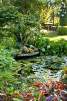 Pond in country garden with reclining sculpture on wooden decking and seating area in background