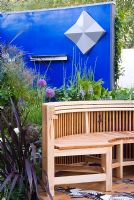 A circular wooden seating area surrounded with planting of mixed perennials, grasses, bulbs and shrubs with a backdrop of a blue painted wall