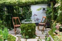 Fantasy moss-covered outdoor room, including perspex table with moss-covered base, plus wooden chairs. Raised beds with moss edging, Irises and dwarf Acer palmatum cultivars. Stone flags on floor with moss growing in cracks - The Green Door, Design - Kazuyuki Ishihara