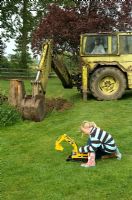 Child playing with toy digger on lawn with real digger digging hole behind - Pannells Ash Farm, West Essex