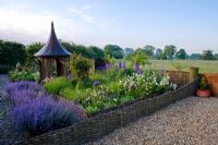 View of flower garden with willow pergola, arbour and fencing - Countryside in background. Planting includes Nepeta racemosa 'Walker's Low' and Persicaria bistorta 'Superba'.