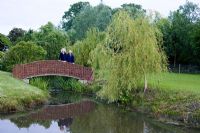 Susie and Robert Yates on willow bridge across pond with Salix x sepulcralis var. chrysocoma and candelabra primulas to right.