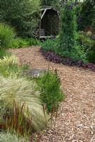 Herbaceous border and ornamental grass border with bark chippings path leading to a wooden arbour with swinging wooden seat - The Rainbow garden, 'Wedgwood', Hesketh Bank, Lancashire 