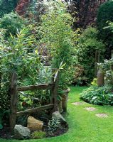 Tall bamboo against old weathered wooden fence with large stones, backed by mature trees and stepping stones in lawn leading to another area of the garden - Courtwood House, Staffordshire NGS