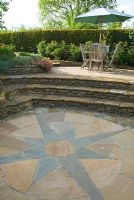Stone terrace with seating area above decorative sunken circular patio, connected by steps and framed with beds of sun loving plants including Roses, Rock Roses, Thymes and Lavender