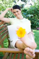 Woman sitting using laptop computer on wooden bench in the garden with yellow flower between her toes