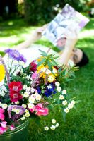 Woman lying on lawn looking at magazine with bucket of flowers in foreground