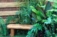 Foliage plants surround a wooden bench with a bamboo and brushwood screen fence in the 'Hideaway' garden, RHS Hampton Court Flower Show