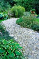 Pebble path with narrow border of shrubs and perennials leading to a seating area beside a pond - Barleywood, Hampshire