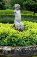 Alchemilla mollis forms a base to a figurative sculpture, with a circular moat of water, hooped railings and lavender in background