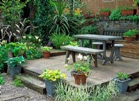Raised dining patio of sandstone, marble table and benches with Hostas in pots and grasses in gravel railway sleeper area in small jungly urban garden planted with exotics - West Park Road, Kew