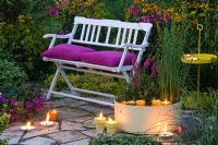 White wooden bench with pink cushions on patio with lit candles, water feature and planting of Lythrum and Scirpus