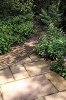 Mixed paving of Indian sandstone and brick in dappled shade, groundcover of Geranium oxonianum and Sylvaticum in borders, Aucuba, buxus and Taxus surrounding