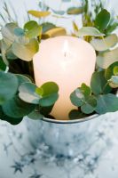 Table decoration of white candle in silver zinc bucket with eucalyptus foliage
