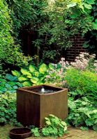 In a small suburban garden, a ceramic tank has been recycled from an old brewery to make a water feature. Surrounded by a planting of Hostas, Asters and Alchemilla mollis.