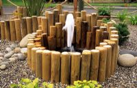 Sunken small water feature with fountain surrounded by lengths of short wooden stakes, gravel and pebbles 