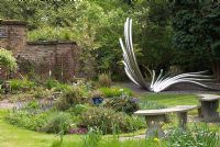 Curved stone benches overlooking small cicular bed in Dave's Garden and 'Nature's Breeze', a stainless steel tubular sculpture created by Sue Sharples, inspired by studies of yucca and cordyline plants - Poulton Hall, NGS garden, Cheshire