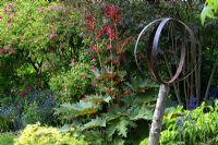 Metal sculpture on plinth backed by mixed border including Rheum and Rosa in May - Ornamental rhubarb and roses