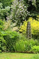 Mixed border of shrubs and perennials including Darmera peltata, Rosa 'Paul's Himalayan Musk' and wooden obelisks