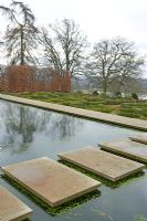 Walkway of concrete paving stones across pond, hornbeam hedge behind and box parterre - Broughton Grange in February 