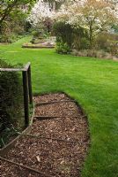 Wooden steps and rail with bark chippings as mulch, leading to lawn and borders with flowering shrubs and trees including Magnolia, Camellia and Amelanchier in Spring at 'Briarfield', Cheshire