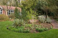 Spring view of secluded garden with mature shrubs, circular central bed, brick wall and gravel path leading to seating area at the back of the bungalow at 'Briarfield' Cheshire