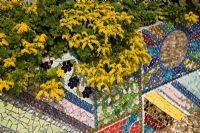 Bright yellow sedums tumble over mosaic covered wall 