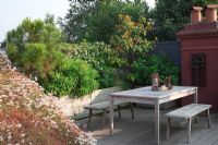 Seating area with wooden table and benches in green corner, planting in large wooden containers, Euphorbia mellifera, Choisya tenata, Magnolia grandiflora, Pinus pinea, Trachleospermum jasminoides and Buxus semperviren cubes with Sedum roof