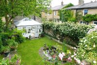 View looking down into sunken garden with conservatory on side of house. Small oval pond, walls covered with rambling and climbing Roses including 'Alberic Barbier' and 'Climbing Iceberg' - New Square, Cambridge