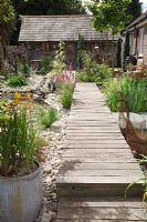 Seaside Inspired garden. Timber decking pathway through seaside garden leading to rustic beach hut. Crocosmia planted in old metal dolly tub, wirework geese sculptures, Lysimachia - Loosestrife and pebble beach.