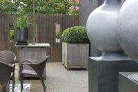 Terrace in summer with large grey urns on plinths. Clipped Buxus - Box in large grey planters in front of black painted wall.

