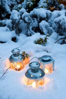 Three tea light candle lanterns set in snow on flower bed