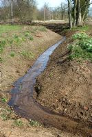 Newly cleaned boundary ditch between bottom of a cottage garden and agricultural land beyond, showing pipe for drainage of garden area into ditch