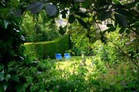 View through foliage to country garden with deck chairs on lawn and a stone sundial backed by a Yew hedge and topiary bird. The Orchards, Litchborough