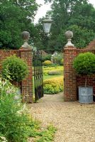 High garden wall with brick pillars, topped with stone balls, standard box trees in lead planters on either side. Open gate leading to well tended lawn area with yellow and gold planting scheme - Frith Hill, Northchapel