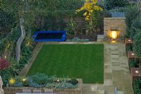 Contemporary urban garden with lighting, lawn, raised brick border, water feature and play area with sunken trampoline - London