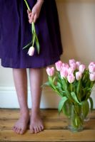 Woman in a dress, standing barefoot alongside a bunch of pink tulips in a vase 