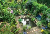 Elevated view of small urban garden with pond, fountain, pot plants, topiary shapes, seating area and Rosa - Rose growing over arch. Hackney, London