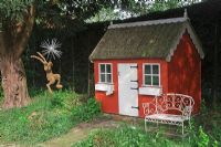 Children's playhouse with thatched roof, wrought iron bench and willow woven hare under shady tree - Northend