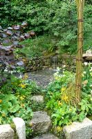 Natural arch above border of Iris, Trollius, Rodgersia and Alchemilla mollis, Corylus maxima 'Purpurea', Primula chungensis. Small waterfall. The 'Music on the Moors' garden - Gold medal winner at RHS Chelsea Flower Show 2010 
 