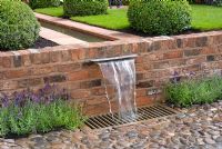 Water flowing along a rill, edged with conical Buxus - Box topiary, and falling over brick wall into drain and cobbled surface. The Russell Watkinson Landscapes 'It's a Reflection of Life' garden - RHS Tatton Flower Show 2010