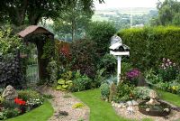Compact corner of small back garden overlooking hills in distance with mixed borders, Leylandii hedge, gravel and cobble path with mat forming plants leading to ornate gate in wooden fence, and features including - a painted bird table, a half barrel water feature with submerged head waterspout and hands, stone gargoyle, clock face made from painted table top and carved stone sculpture with 'Croeso' - Welcome in Welsh at 'Trevinia', Stubbins, Lancashire NGS
 